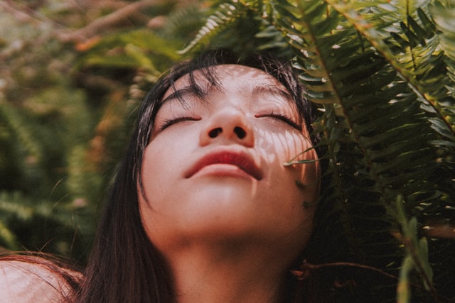 A young asian women tilts her head back with eyes closed in a tropical forest. She seems at peace.